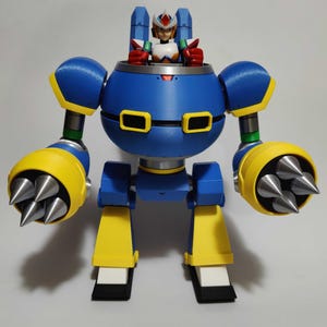 May include: A blue, yellow, and white robot toy with a figure inside. The robot has large, round arms with silver spikes and yellow accents. The figure is wearing a white, red, and blue suit. The toy is standing on a white surface.