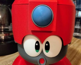 May include: A 3D-printed, red, cartoon-like container with large white eyes, green pupils, and a red mouth. The container has a blue circle on the front and is supported by three red feet. The container is sitting on a wooden surface.