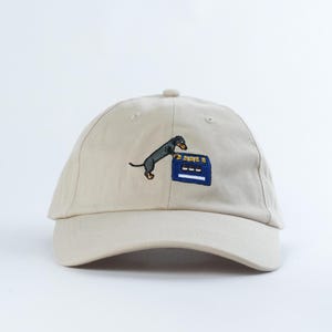 May include: A beige baseball cap featuring an embroidered graphic of a black and tan dachshund standing on its hind legs, reaching towards a blue crate filled with yellow and white bottles. The cap has a curved brim and a structured crown.