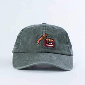 Könnte beinhalten: Eine dunkelgrüne Baseballkappe mit einer Stickerei eines braunen Dackels, der auf seinen Hinterbeinen neben einem roten rechteckigen Objekt mit gelben und weißen Details steht. Die Kappe hat einen gebogenen Schirm und eine Krone.