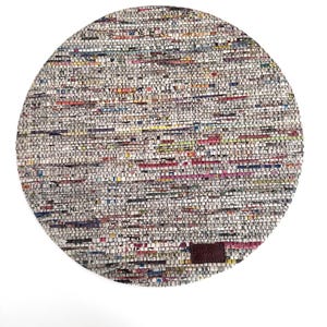 May include: A circular placemat constructed from tightly woven, multicolored strips of recycled paper or fabric. The surface features a dense, variegated pattern of small rectangular segments in shades of white, gray, red, blue, and yellow, with a brown leather tag.