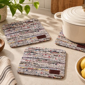 May include: Three square trivets made of woven multicolored fabric strips with a textured, speckled appearance. Each trivet features a small, rectangular brown leather-like patch in the corner. The trivets have a flat, flexible construction.