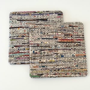 May include: Two square coasters made from woven fabric scraps. The coasters have a neutral color palette with flecks of red, blue, and green. The texture of the coasters is visible, suggesting a handmade or upcycled product.