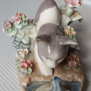 May include: A porcelain figurine of a grey and white cat with a black tail, crouching near a blue frog. The cat is surrounded by pink and white flowers and green leaves.
