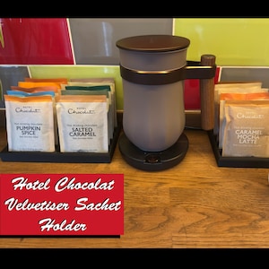 May include: A grey Hotel Chocolat Velvetiser with a wooden handle and a brown lid. The image displays hot chocolate sachets in flavours such as Pumpkin Spice, Salted Caramel, and Caramel Mocha Latte, plus a white mug. The text reads 'Hotel Chocolat Velvetiser Sachet Holder'.