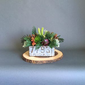 May include: A floral arrangement in a rectangular metal container with the numbers "7793" and the word "FLORIDA" on it. The arrangement includes green, orange, and purple faux flowers and greenery, set on a wooden slice.