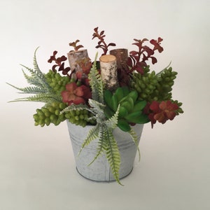 May include: A rustic succulent arrangement in a galvanised metal bucket. The arrangement features a variety of green succulents, red-brown leaves, and birch wood accents.
