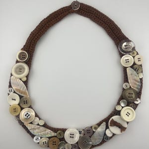 以下が含まれることがあります： 白、茶色、銀色のボタンが飾られた、茶色の手編みネックレス。 ボタンは様々なサイズと質感で、ユニークで手作りのデザインを作り出しています。 このネックレスは、存在感のあるアクセサリーです。