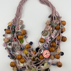 May include: A multi-strand necklace made of light pink crocheted yarn, featuring an assortment of attached buttons in various shapes, sizes, and colors, including wooden beads, floral patterns, and a metal toggle clasp at the top.