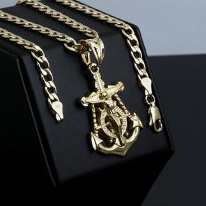 Op de afbeelding: Goudkleurige ketting met een gedetailleerde ankerhanger met een religieuze figuur. De hanger hangt aan een dikke, gouden ketting met een karabijnsluiting. De sieraden worden gepresenteerd op een zwart oppervlak.