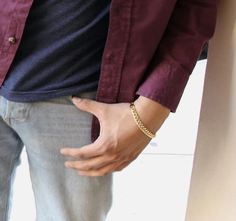 May include: A gold curb chain bracelet is worn on a person's wrist. The bracelet is a bright gold color and has a classic, linked design. The person is wearing a burgundy button-down shirt and blue jeans.