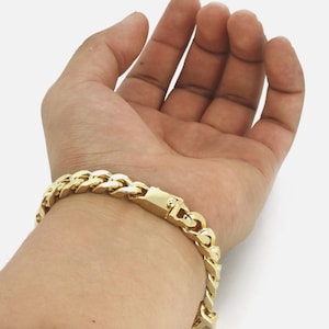 May include: A gold-colored curb chain bracelet. The bracelet is made of interlocking links and features a secure clasp. The jewelry is worn on the wrist and is displayed against a light background.