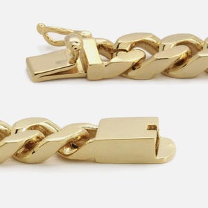 May include: Close-up of a gold chain bracelet with a box clasp. The links are thick and rounded, with a polished finish. The clasp is rectangular with a secure closure.