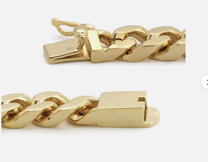 May include: Close-up of a gold chain bracelet with a box clasp. The links are thick and rounded, with a polished finish. The clasp is rectangular with a secure closure.