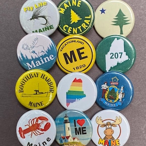 May include: A collection of twelve round Maine-themed buttons. Designs include a moose, a lobster, a lighthouse, a rainbow-colored state outline, and text such as "Fly Life", "Maine Central", and "I Love ME".