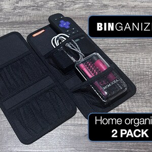 May include: A black fabric organizer with multiple pockets and mesh compartments. It is open and filled with items like a remote control, cords, and a pink case. The text "BINGANIZER" and "Home organizer 2 PACK" is visible on the image.