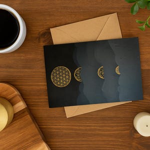 May include: A still-life image featuring a cup of coffee, stationery, and candles on a wooden surface. The stationery includes a black card with a gold geometric design and a brown envelope. Two gold coasters and two white candles are also present.
