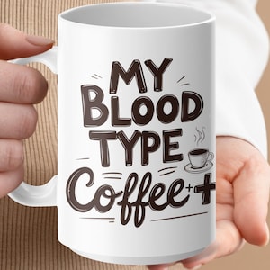 Puede incluir: Taza de café de cerámica blanca con el texto "MY BLOOD TYPE Coffee +" en letras de color marrón oscuro. También se incluye un pequeño gráfico de taza de café. La taza la sostiene una persona.
