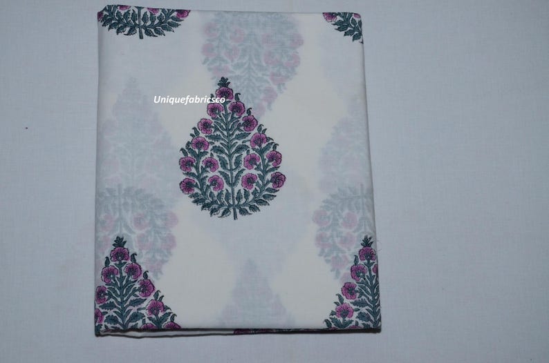 May include: A folded white fabric featuring a repeating pattern of dark green leafy stems with pink floral buds. The text "Uniquefabricsco" is printed in white on the central floral motif. The fabric is rectangular and neatly folded.