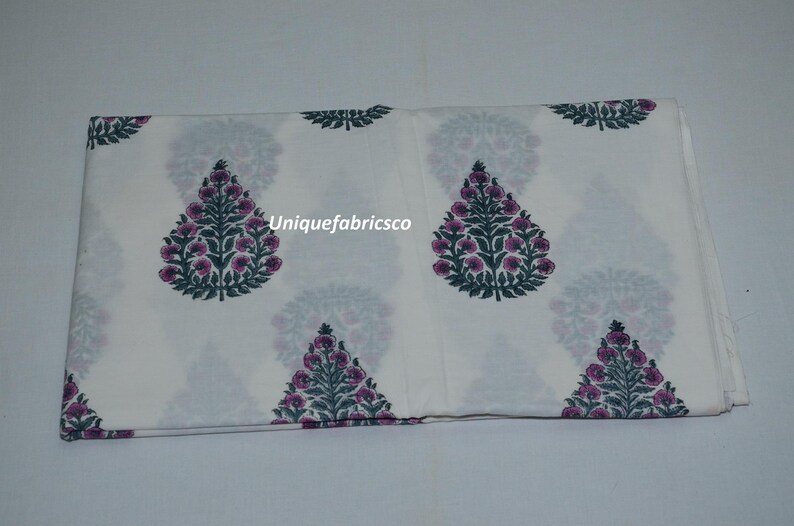 May include: A folded piece of white fabric featuring a repeating pattern of dark green and purple floral tree motifs. The text "Uniquefabricsco" is printed in white across the center of the fabric, which displays a symmetrical botanical design.