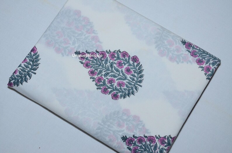 May include: A folded piece of white fabric featuring a repeating pattern of teardrop-shaped floral motifs in muted green and pink. The text "Uniquefabricsco" is printed in small white letters near the center of the fabric surface.
