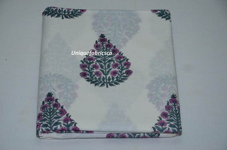 May include: A folded piece of white fabric featuring a repeating pattern of dark green leafy trees with pink flowers. The text "Uniquefabricsco" is printed in white across the center of the fabric. The design consists of symmetrical floral motifs.