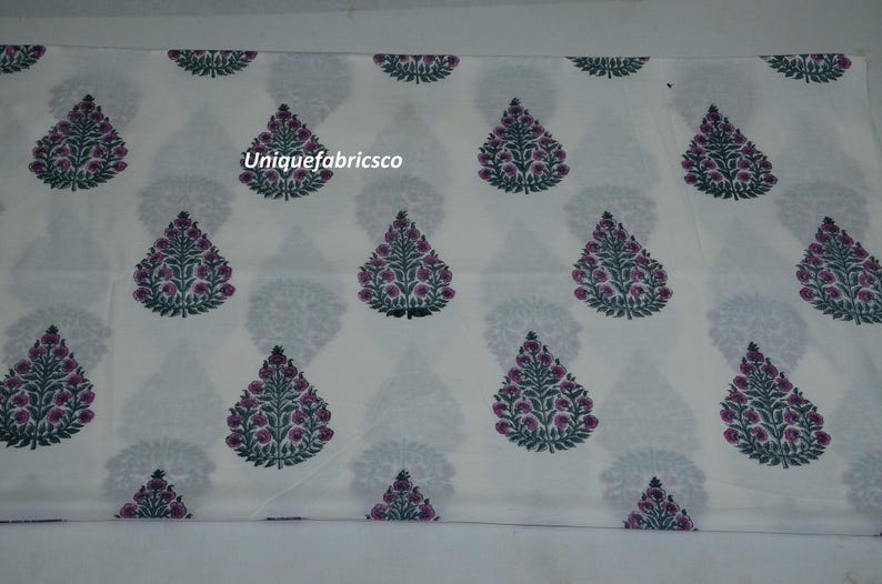 May include: A white fabric featuring a repeating pattern of teardrop-shaped floral motifs in dark green and magenta. The design includes the text Uniquefabricsco printed in white over one of the central floral elements on the textile surface.