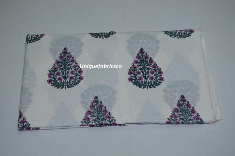 May include: A folded piece of white fabric featuring a repeating pattern of dark green and purple floral tree motifs. The text "Uniquefabricsco" is printed in white across the center of the fabric, which displays a symmetrical botanical design.