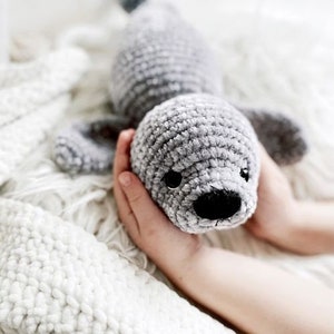 May include: A gray crocheted seal plush toy with black eyes and a black nose. The seal is being held in the hands of a person.
