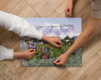Può includere: Due persone che lavorano insieme per completare un puzzle di una catena montuosa con fiori selvatici in primo piano. Il puzzle è intitolato "Mount Rainier, Washington."
