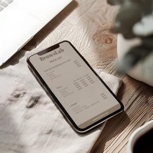 May include: A smartphone displaying a price list from BrowsLab, with categories for eyelash extensions, fills, and lashes. The phone is on a wooden surface, near a laptop and a cup of coffee.