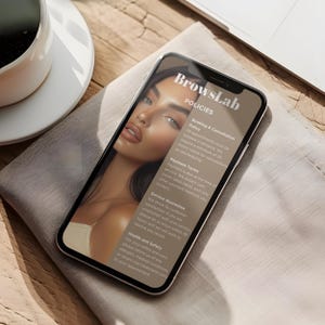 May include: A smartphone displays the "BrowsLab" policies, including booking, payment, and service guarantee details. The screen shows a portrait of a woman with makeup. The phone rests on a neutral-toned cloth, with a coffee cup and a laptop visible.