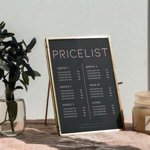 May include: A framed black pricelist with gold trim stands on a pink surface. The text "PRICELIST" is in pink, with service descriptions below. A glass vase with greenery and a small jar with a label are also visible.