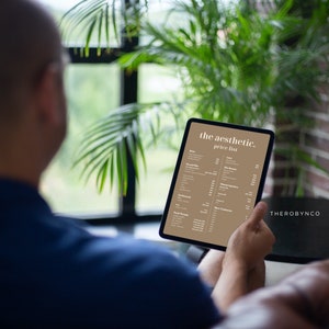 May include: A person holding a tablet displaying "the aesthetic. price list" in a tan and white color scheme. The tablet is held in front of a blurred background of a window, plants, and brick wall.