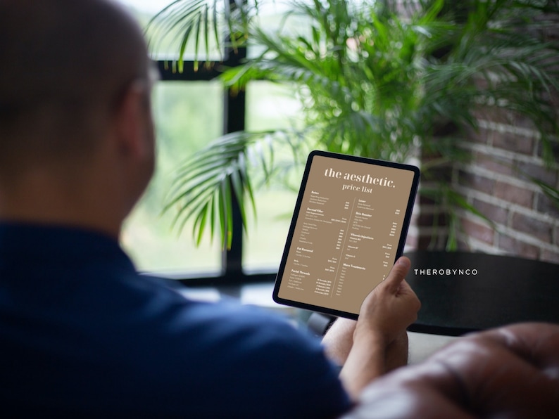 May include: A person holding a tablet displaying "the aesthetic. price list" in a tan and white color scheme. The tablet is held in front of a blurred background of a window, plants, and brick wall.