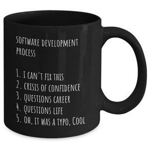 May include: Black coffee mug with white text that reads "SOFTWARE DEVELOPMENT PROCESS" and a numbered list: "1. I CAN'T FIX THIS, 2. CRISIS OF CONFIDENCE, 3. QUESTIONS CAREER, 4. QUESTIONS LIFE, 5. OH, IT WAS A TYPO, COOL"