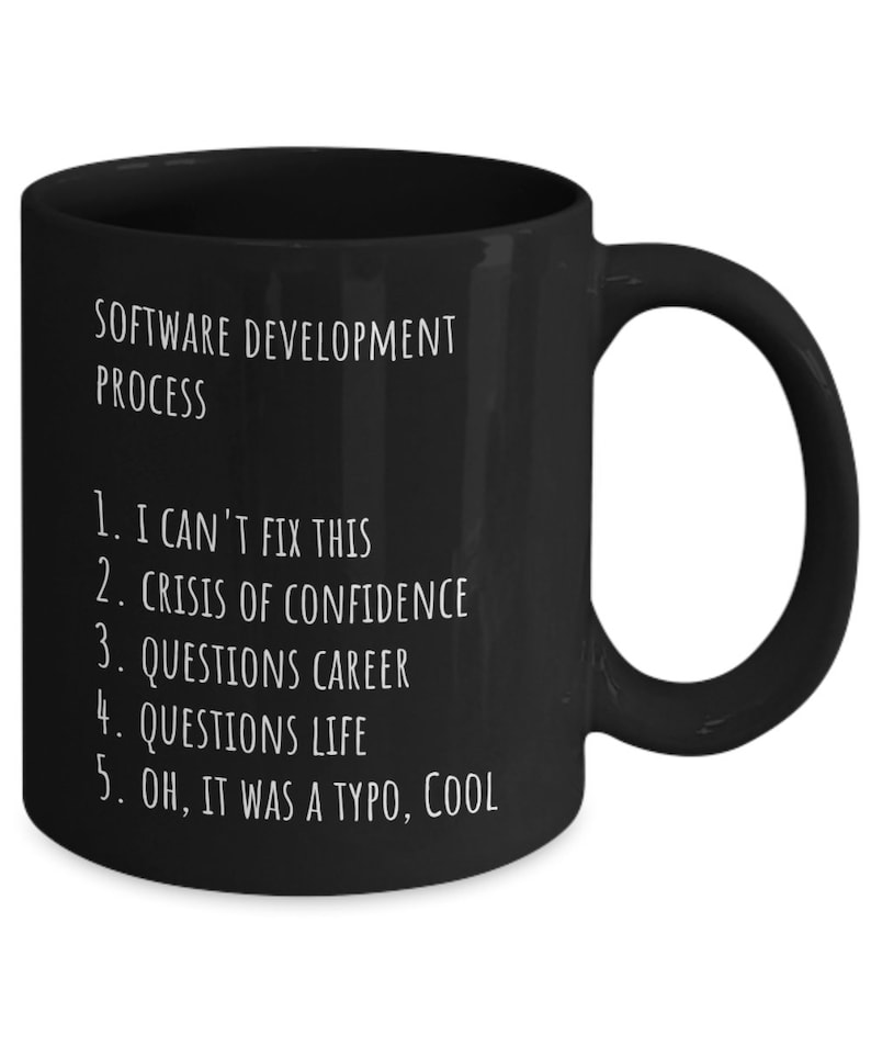 May include: Black coffee mug with white text that reads "SOFTWARE DEVELOPMENT PROCESS" and a numbered list: "1. I CAN'T FIX THIS, 2. CRISIS OF CONFIDENCE, 3. QUESTIONS CAREER, 4. QUESTIONS LIFE, 5. OH, IT WAS A TYPO, COOL"