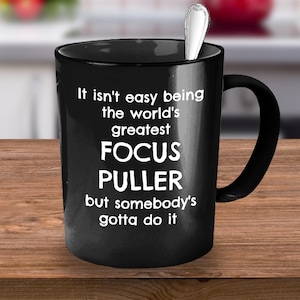 May include: Black coffee mug with white text that reads "It isn't easy being the world's greatest FOCUS PULLER but somebody's gotta do it".
