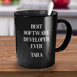 Puede incluir: Una taza de cerámica negra con el texto "BEST SOFTWARE DEVELOPER EVER TARA" en blanco. Una cuchara plateada está dentro de la taza. La taza está sobre una superficie de madera.