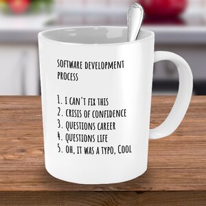 May include: White ceramic coffee mug with black text that reads "SOFTWARE DEVELOPMENT PROCESS" and a numbered list: "1. I CAN'T FIX THIS", "2. CRISIS OF CONFIDENCE", "3. QUESTIONS CAREER", "4. QUESTIONS LIFE", "5. OH, IT WAS A TYPO, COOL".