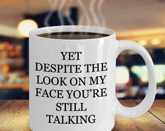 May include: White ceramic coffee mug with black text that reads "YET DESPITE THE LOOK ON MY FACE YOU'RE STILL TALKING".