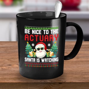 Może przedstawiać: Czarny ceramiczny kubek z napisem "BE NICE TO THE ACTUARY SANTA IS WATCHING" w kolorze białym, czerwonym i zielonym. Kubek przedstawia grafikę Świętego Mikołaja w okularach przeciwsłonecznych i czapce Mikołaja, z choinkami i prezentami.
