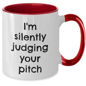 Puede incluir: Taza de café de cerámica blanca con borde y asa rojos. La taza tiene texto negro que dice "Estoy juzgando silenciosamente tu discurso."