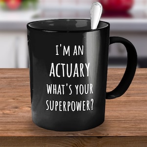 Może przedstawiać: Czarny ceramiczny kubek z białym napisem "I'M AN ACTUARY WHAT'S YOUR SUPERPOWER?". Srebrna łyżeczka spoczywa wewnątrz kubka. Kubek stoi na drewnianej powierzchni.
