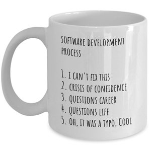 May include: White ceramic coffee mug with black text that reads "SOFTWARE DEVELOPMENT PROCESS" and a numbered list: "1. I CAN'T FIX THIS 2. CRISIS OF CONFIDENCE 3. QUESTIONS CAREER 4. QUESTIONS LIFE 5. OH, IT WAS A TYPO, COOL"