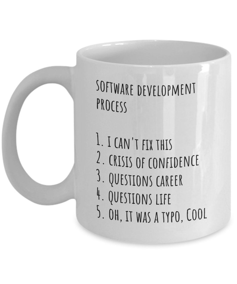 May include: White ceramic coffee mug with black text that reads "SOFTWARE DEVELOPMENT PROCESS" and a numbered list: "1. I CAN'T FIX THIS 2. CRISIS OF CONFIDENCE 3. QUESTIONS CAREER 4. QUESTIONS LIFE 5. OH, IT WAS A TYPO, COOL"
