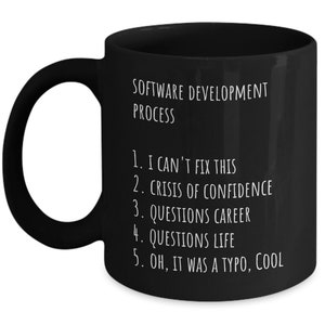 May include: Black coffee mug with white text that reads "SOFTWARE DEVELOPMENT PROCESS" and a numbered list: "1. I CAN'T FIX THIS 2. CRISIS OF CONFIDENCE 3. QUESTIONS CAREER 4. QUESTIONS LIFE 5. OH, IT WAS A TYPO, COOL"
