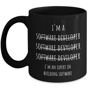 Op de afbeelding: Zwarte keramische mok met witte tekst: "I'M A SOFTWARE DIVELOPER SOFTWARE DEVYLOPER SOFTWARE DEVILOPER I'M AN EXPERT IN BUILDING SOFTWARE". De mok heeft een groot oor.