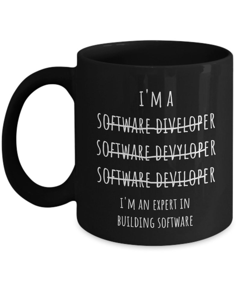 Op de afbeelding: Zwarte keramische mok met witte tekst: "I'M A SOFTWARE DIVELOPER SOFTWARE DEVYLOPER SOFTWARE DEVILOPER I'M AN EXPERT IN BUILDING SOFTWARE". De mok heeft een groot oor.