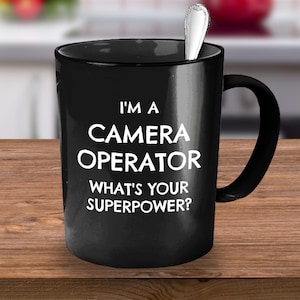 May include: Black coffee mug with white text that reads "I'm a Camera Operator What's Your Superpower?"